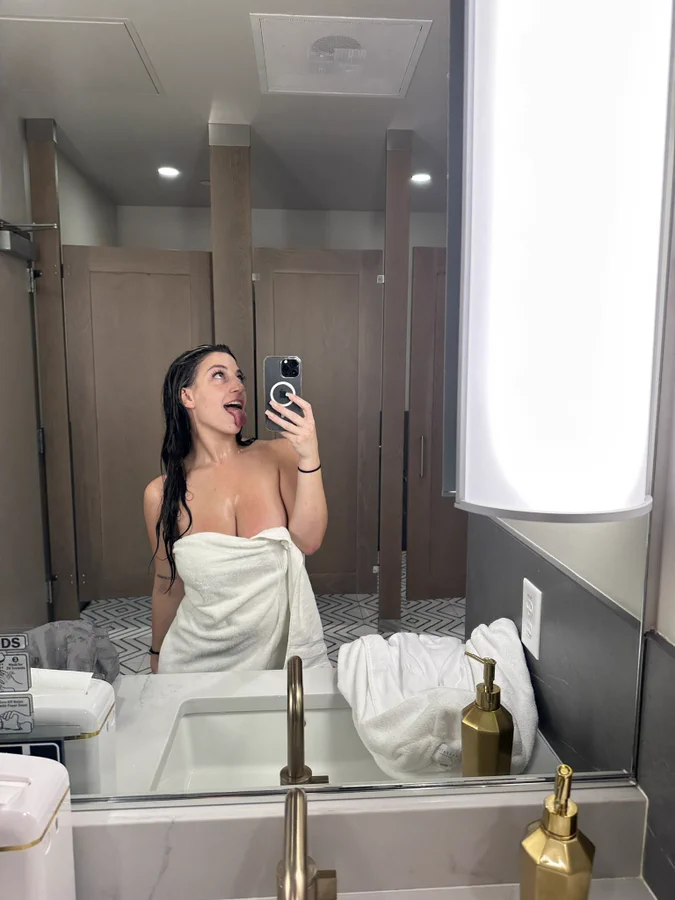 Megan Marie @megmariiee parada con una toalla en el baño sosteniendo un teléfono en una selfie en el espejo