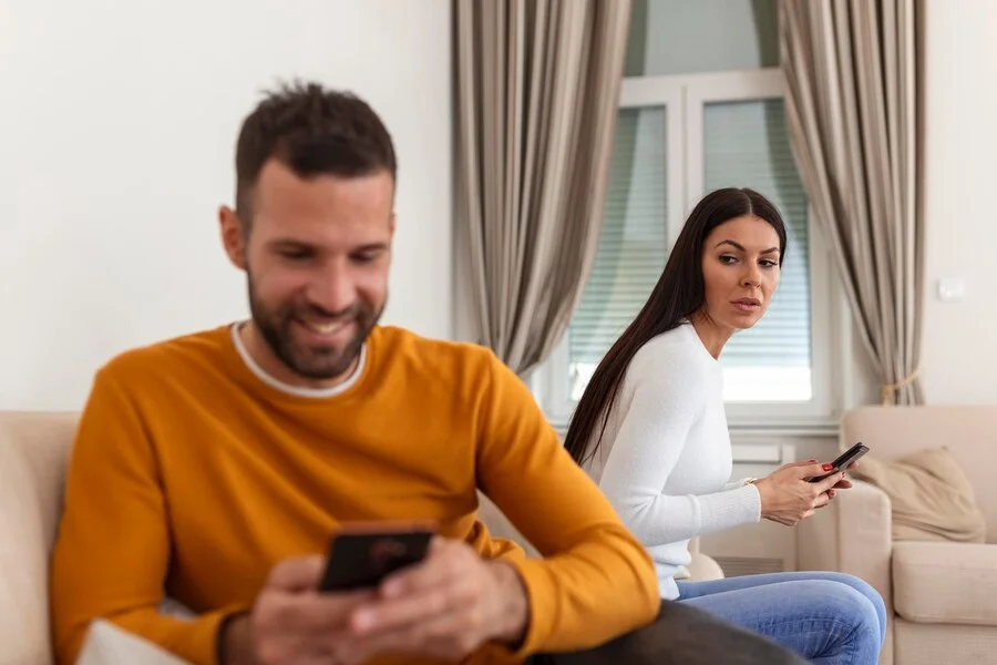 a man is using his phone while smiling while the woman is looking at him worried. it is a picture in a topic of cheating onlyfans